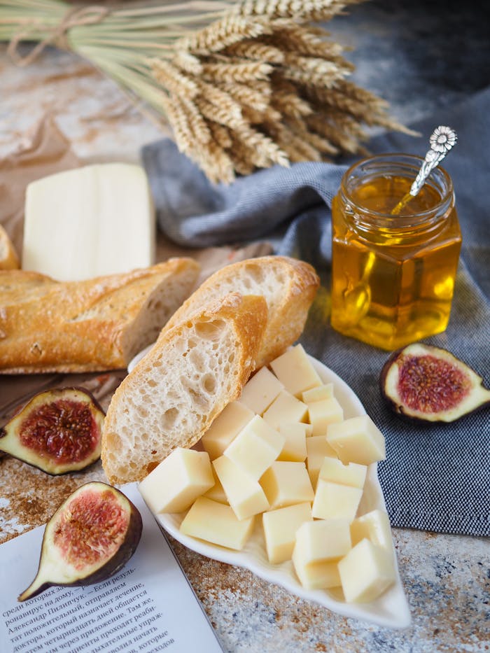 A rustic still life featuring cheese, baguette, figs, and honey on a textured background.