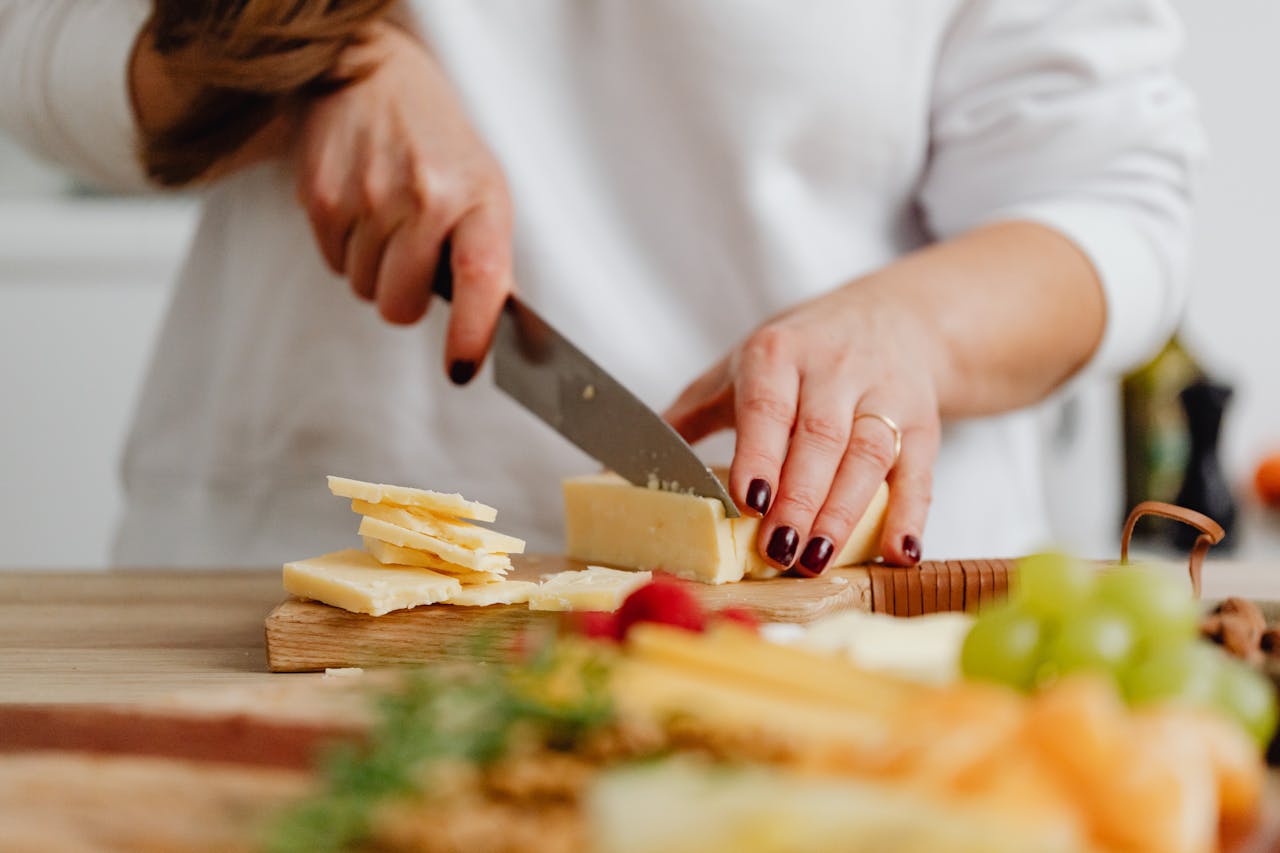 Close-up of hands slicing cheese with fresh fruits in the background, showcasing food preparation.